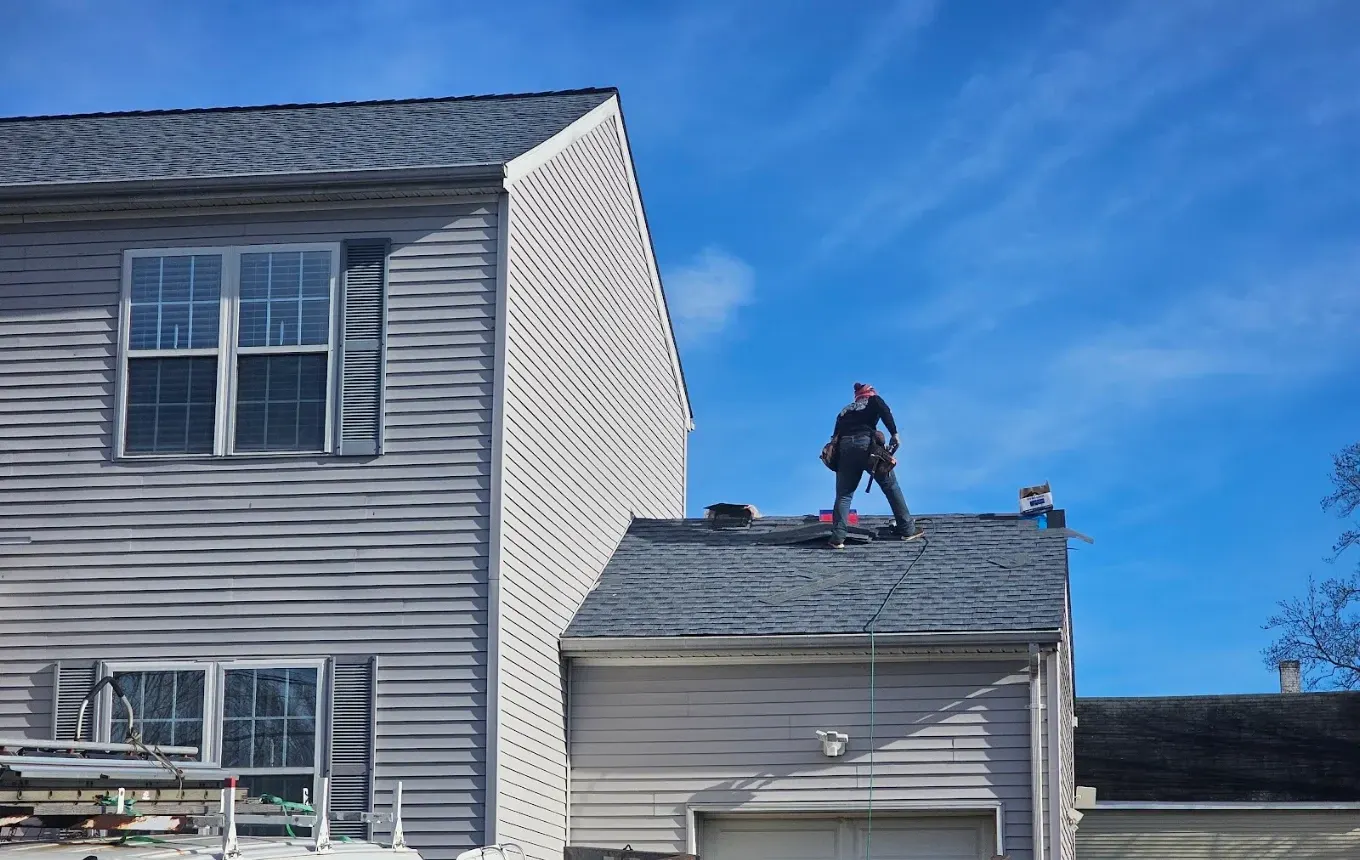 A roofer in a tool belt standing on a garage roof mid tear-off, with a work van and ladder rack parked below and clear blue sky behind.