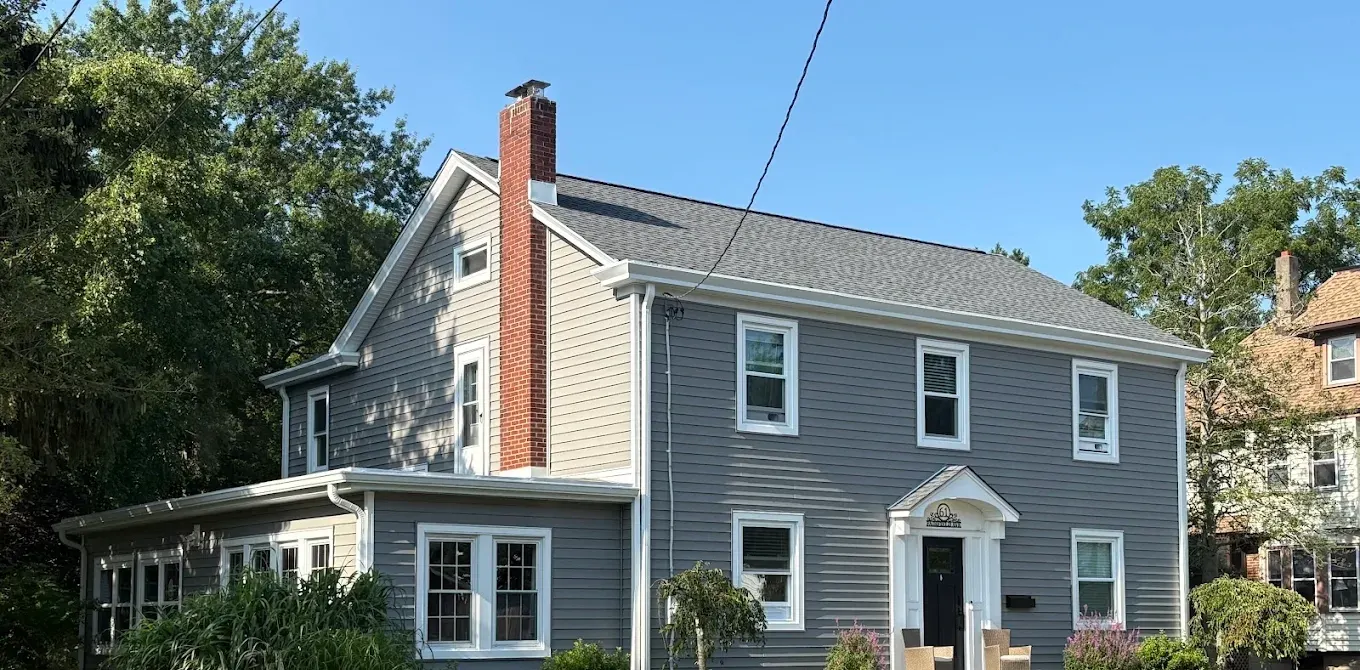 A grey clapboard colonial house seen from the street with a new charcoal shingle roof, white trim and a red brick chimney.
